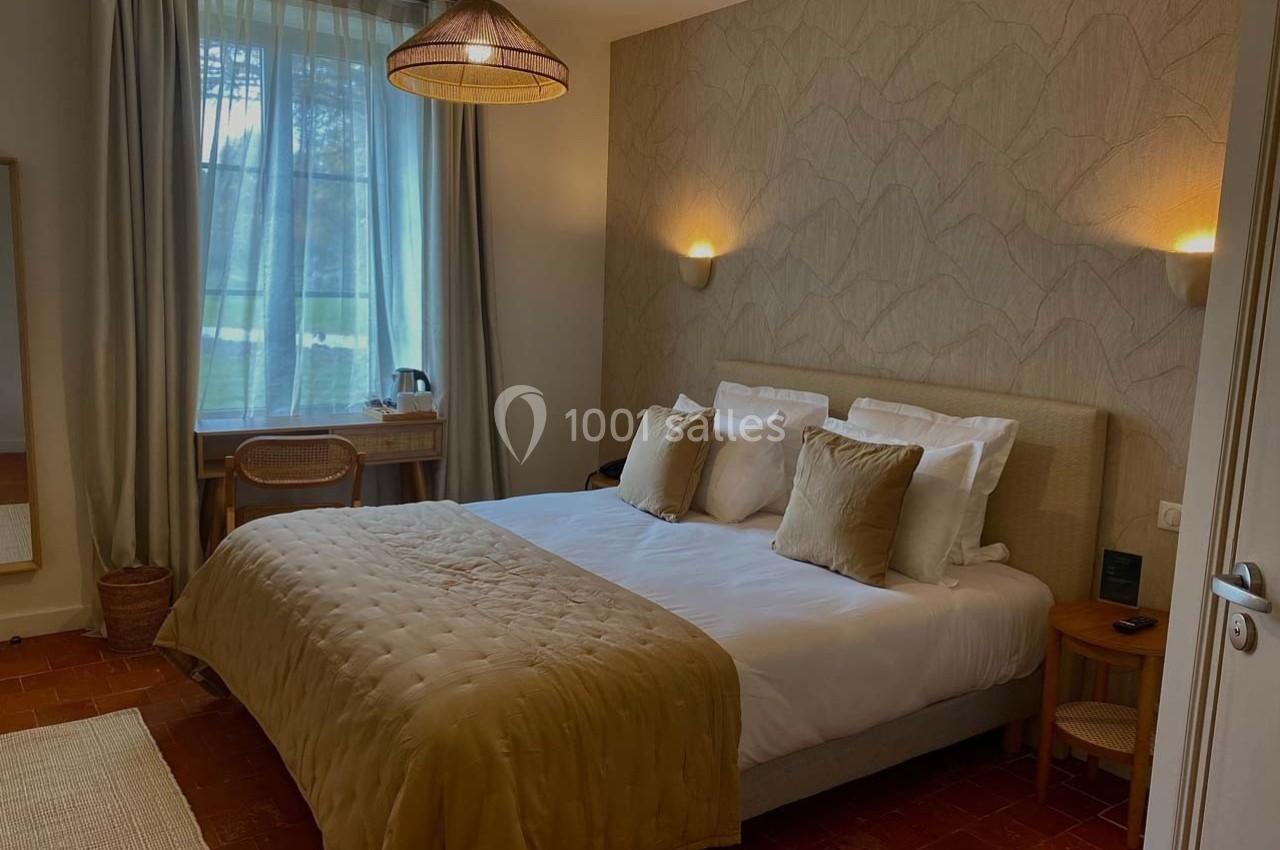 Chambre lumineuse avec lit double, couvre-lit beige, bureau en bois près d'une fenêtre et éclairage mural doux.