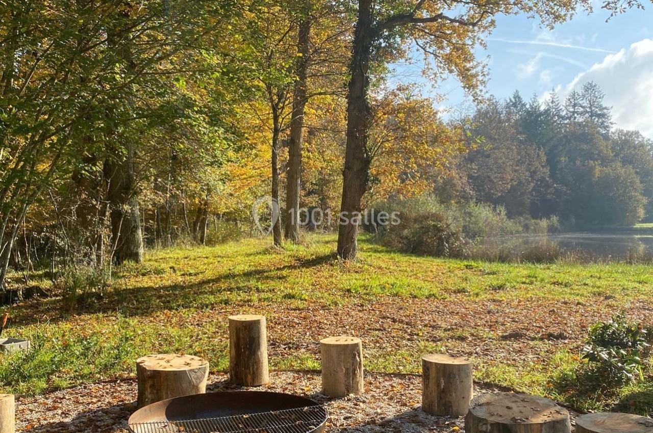 Clairière ensoleillée avec des troncs d'arbres disposés autour d'un foyer, près d'un étang bordé de forêt.