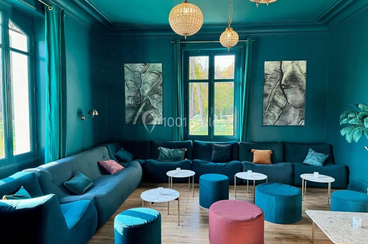 Salon spacieux avec murs et mobilier bleu, canapés en angle, tables basses rondes et suspensions lumineuses.