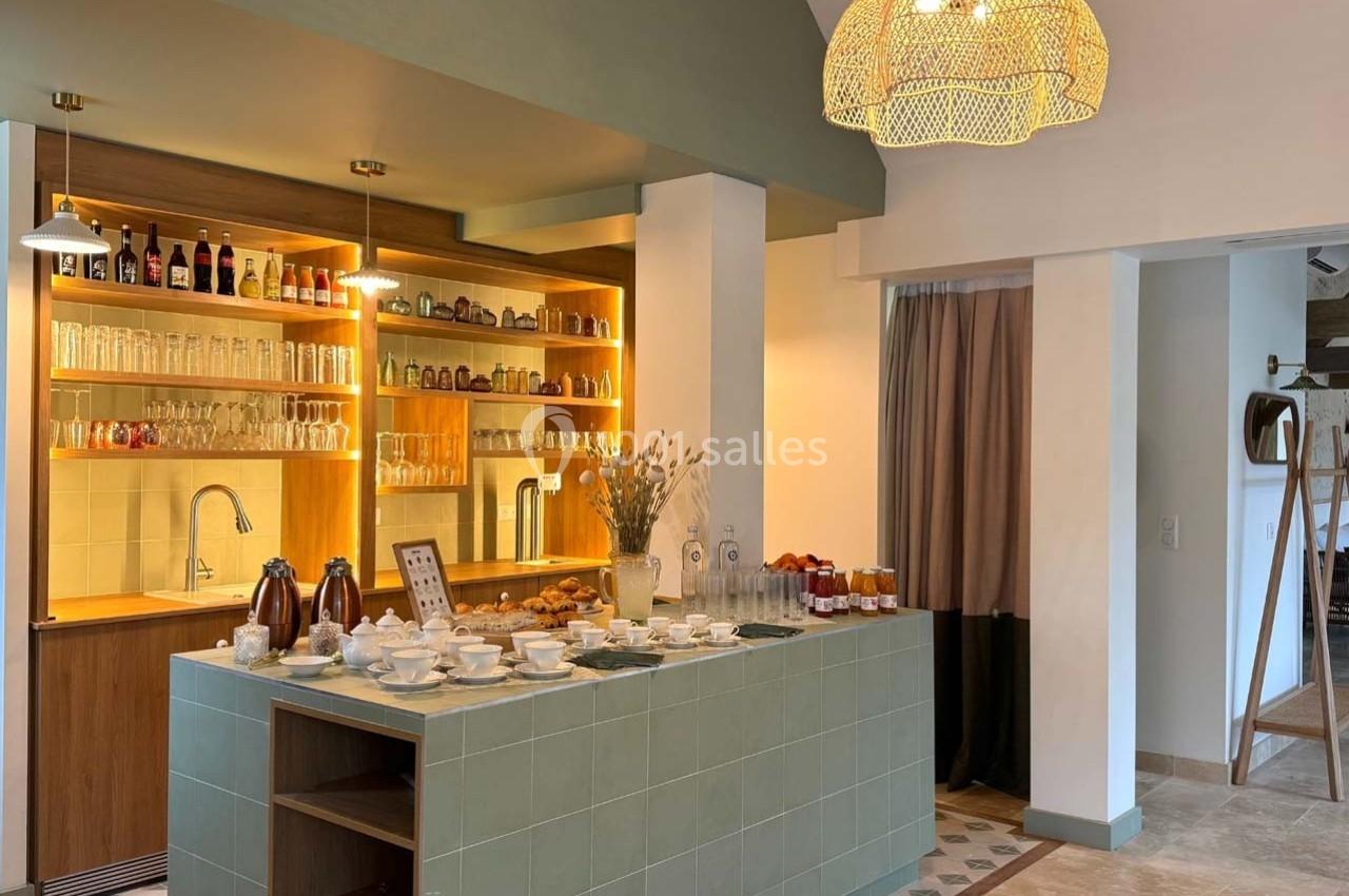 Buffet avec boissons chaudes, viennoiseries et confitures, devant un bar éclairé avec étagères de verres et bouteilles.