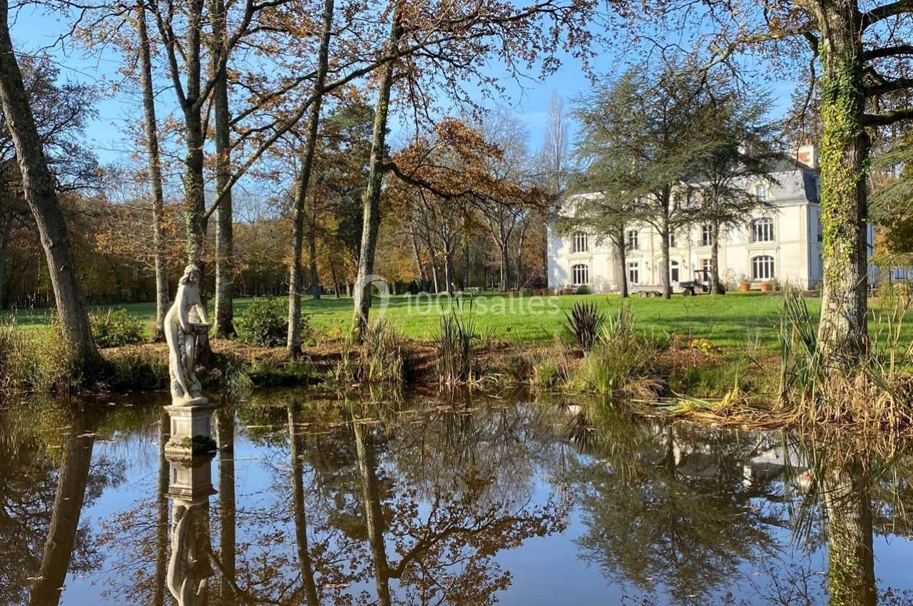 Étang entouré d'arbres avec une statue en pierre au premier plan et un manoir blanc en arrière-plan.