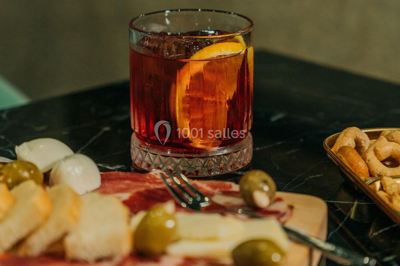 Verre de cocktail rouge garni d'une tranche d'orange, accompagné d'un plateau de charcuterie, fromage et olives.