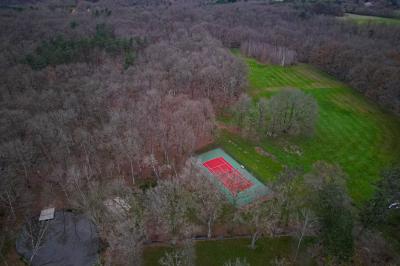 Vue aérienne d'un terrain de tennis rouge entouré de forêt, avec une petite mare et des prairies avoisinantes.
