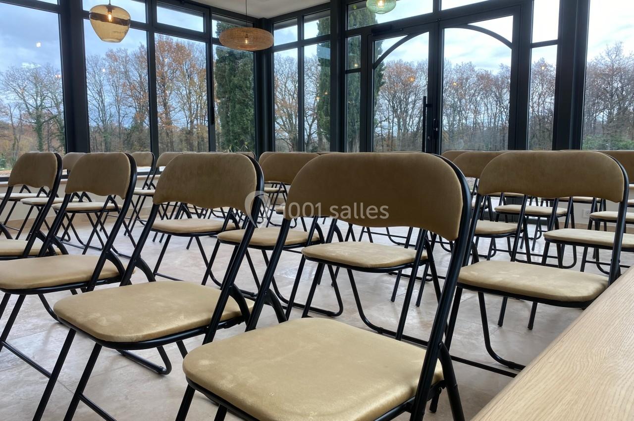 Chaises pliantes beige disposées en rangées dans une salle lumineuse avec de grandes baies vitrées donnant sur des arbres.
