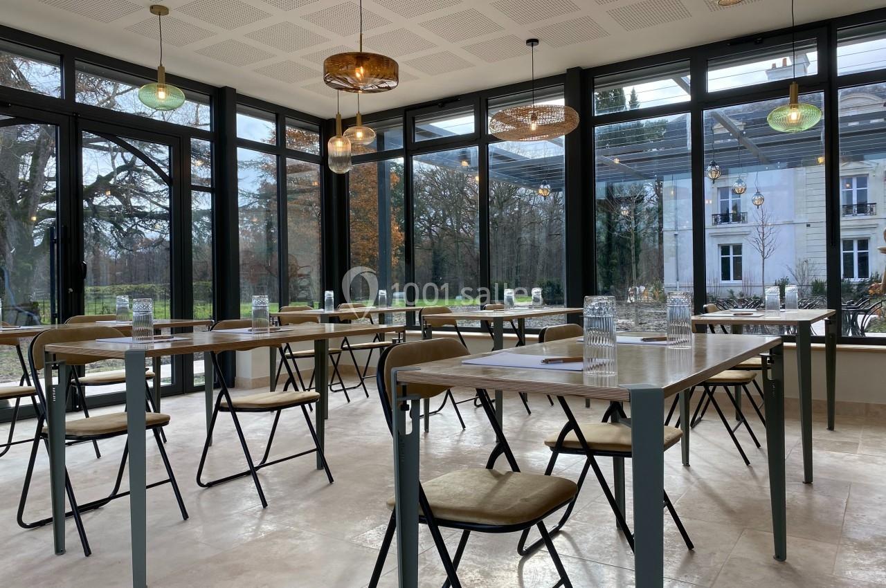 Salle lumineuse avec de grandes baies vitrées, tables et chaises disposées pour une réunion, vue sur un jardin arboré.