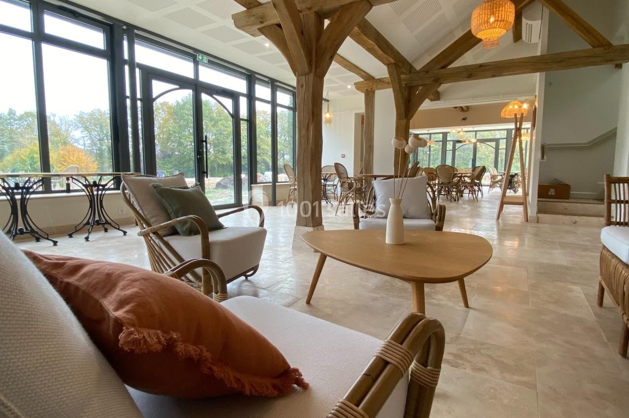 Salle lumineuse avec grandes baies vitrées, poutres en bois, mobilier en rotin et décoration chaleureuse.