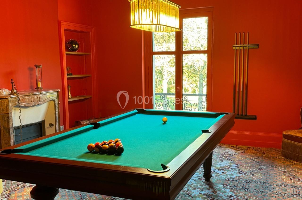 Table de billard avec billes alignées, dans une pièce rouge avec cheminée, étagères et lustre suspendu.