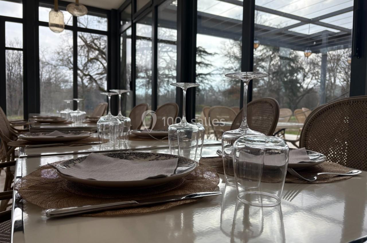 Salle de restaurant lumineuse avec tables dressées, nappes en osier, verres et couverts, donnant sur un extérieur arboré.