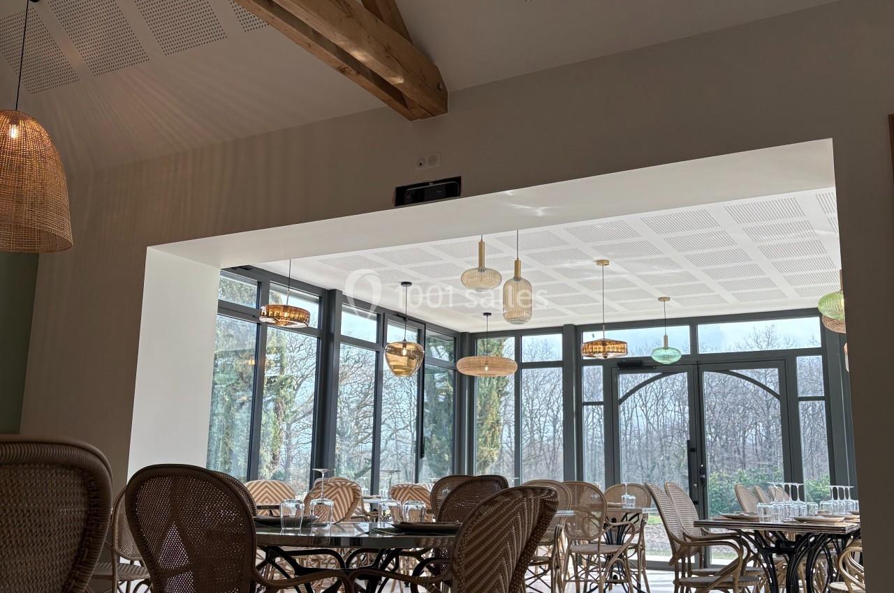 Salle lumineuse avec grandes baies vitrées, mobilier en rotin et plafonniers en osier, vue sur un paysage arboré.