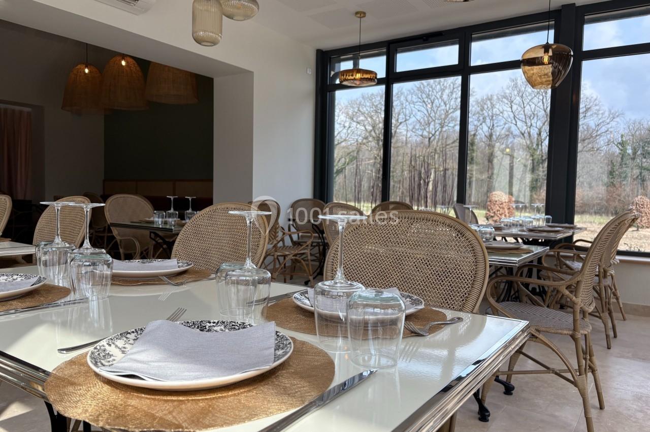 Salle de restaurant lumineuse avec tables dressées, chaises en rotin et grandes baies vitrées donnant sur un paysage boisé.