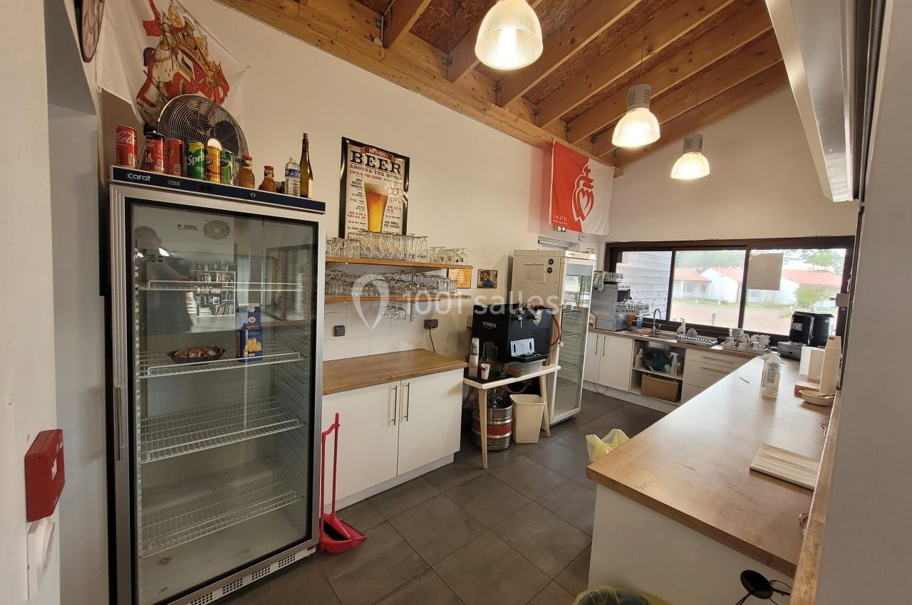 Cuisine équipée avec réfrigérateur, étagères, machine à café et comptoir, éclairée par des suspensions et une fenêtre.