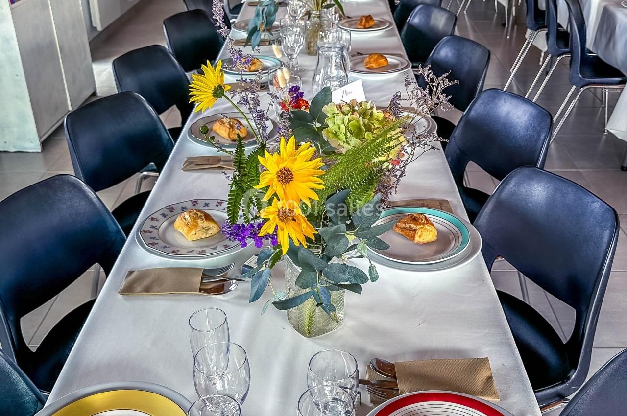 Table dressée avec nappes blanches, assiettes, verres, croissants et bouquets de fleurs colorées dans une salle lumineuse.