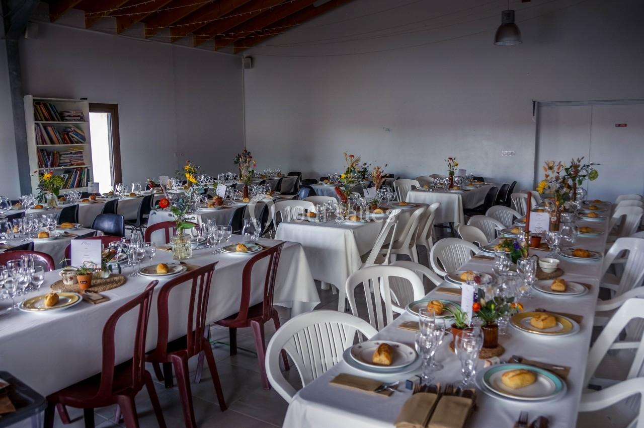 Salle décorée pour un repas avec des tables dressées, nappes blanches, fleurs et couverts disposés.