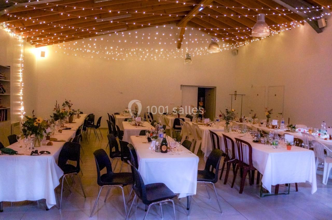 Salle décorée pour un événement, avec tables dressées, nappes blanches, chaises variées et guirlandes lumineuses suspendues.