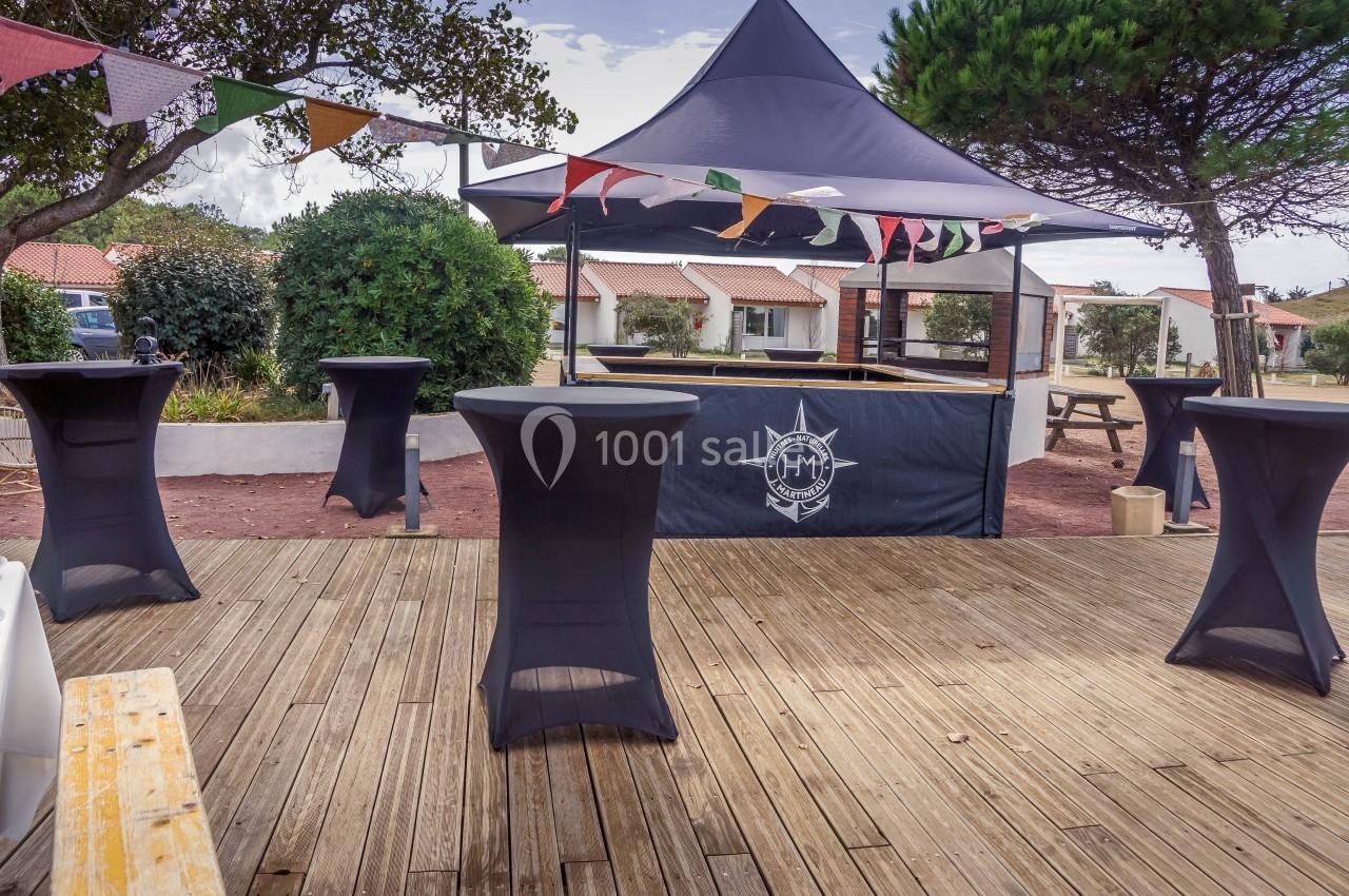 Espace extérieur avec tables hautes noires, guirlandes colorées et tente sous des arbres, sur une terrasse en bois.
