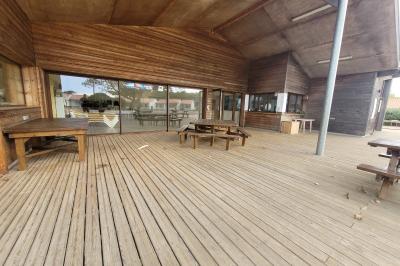 Terrasse en bois avec tables, bancs et grande baie vitrée donnant sur un espace extérieur avec quelques bâtiments au loin.