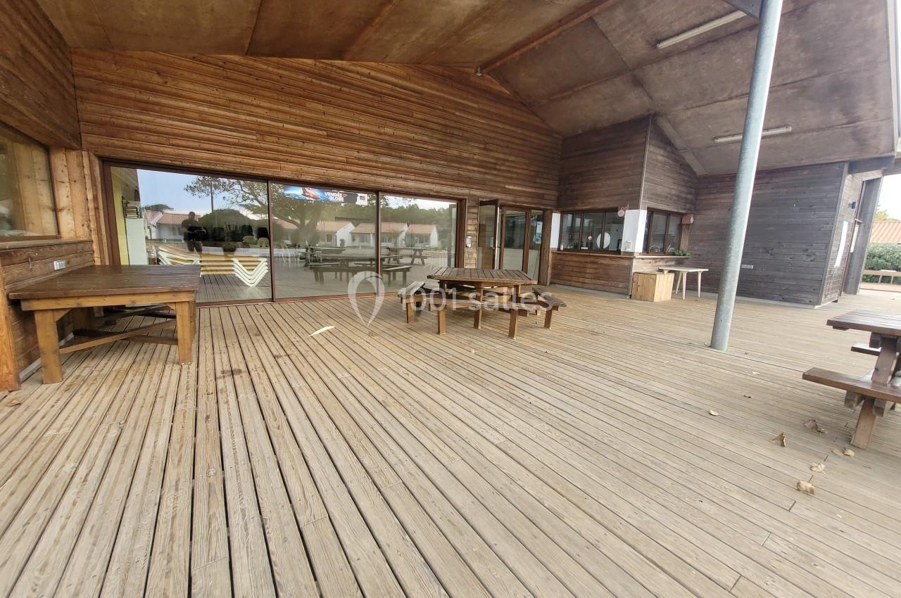 Terrasse en bois avec tables, bancs et grande baie vitrée donnant sur un espace extérieur avec quelques bâtiments au loin.