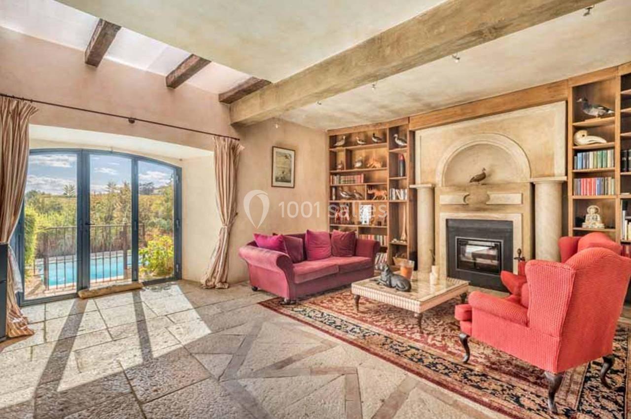 Salon lumineux avec poutres apparentes, cheminée, fauteuils rouges, bibliothèque et vue sur une piscine extérieure.