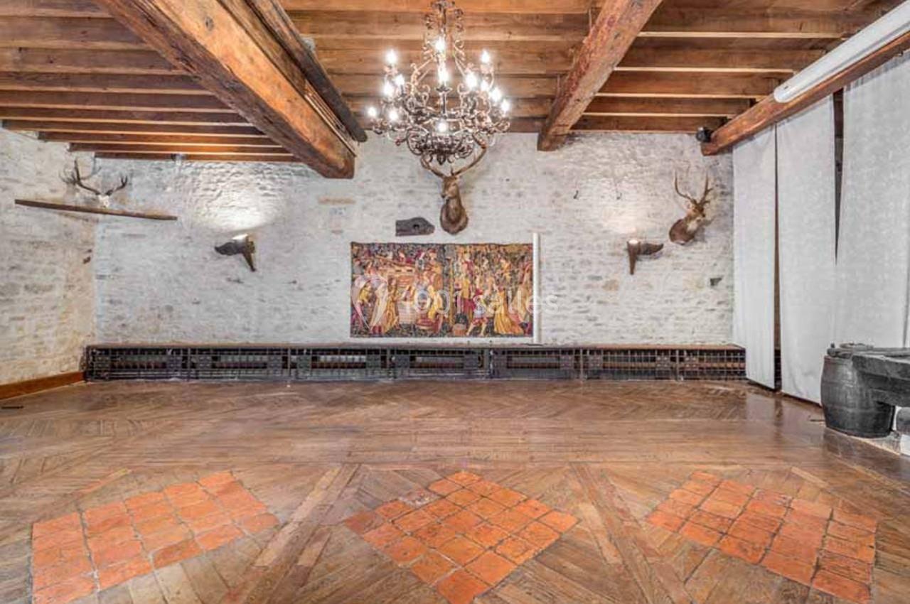 Salle avec murs en pierre, plafond en poutres apparentes, lustre central, tapisseries murales et parquet en bois.
