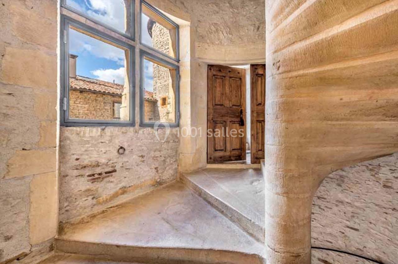 Escalier en pierre en colimaçon menant à une pièce lumineuse avec des murs en pierre et des fenêtres à carreaux.