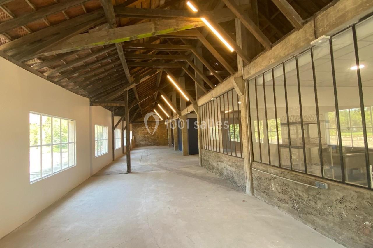 Intérieur d'un bâtiment rénové avec charpente en bois apparente, murs blancs et grandes fenêtres vitrées.