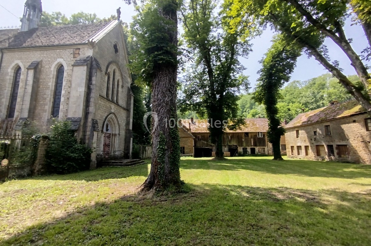Vue d'une petite église en pierre entourée d'arbres et de bâtiments anciens dans un cadre verdoyant.