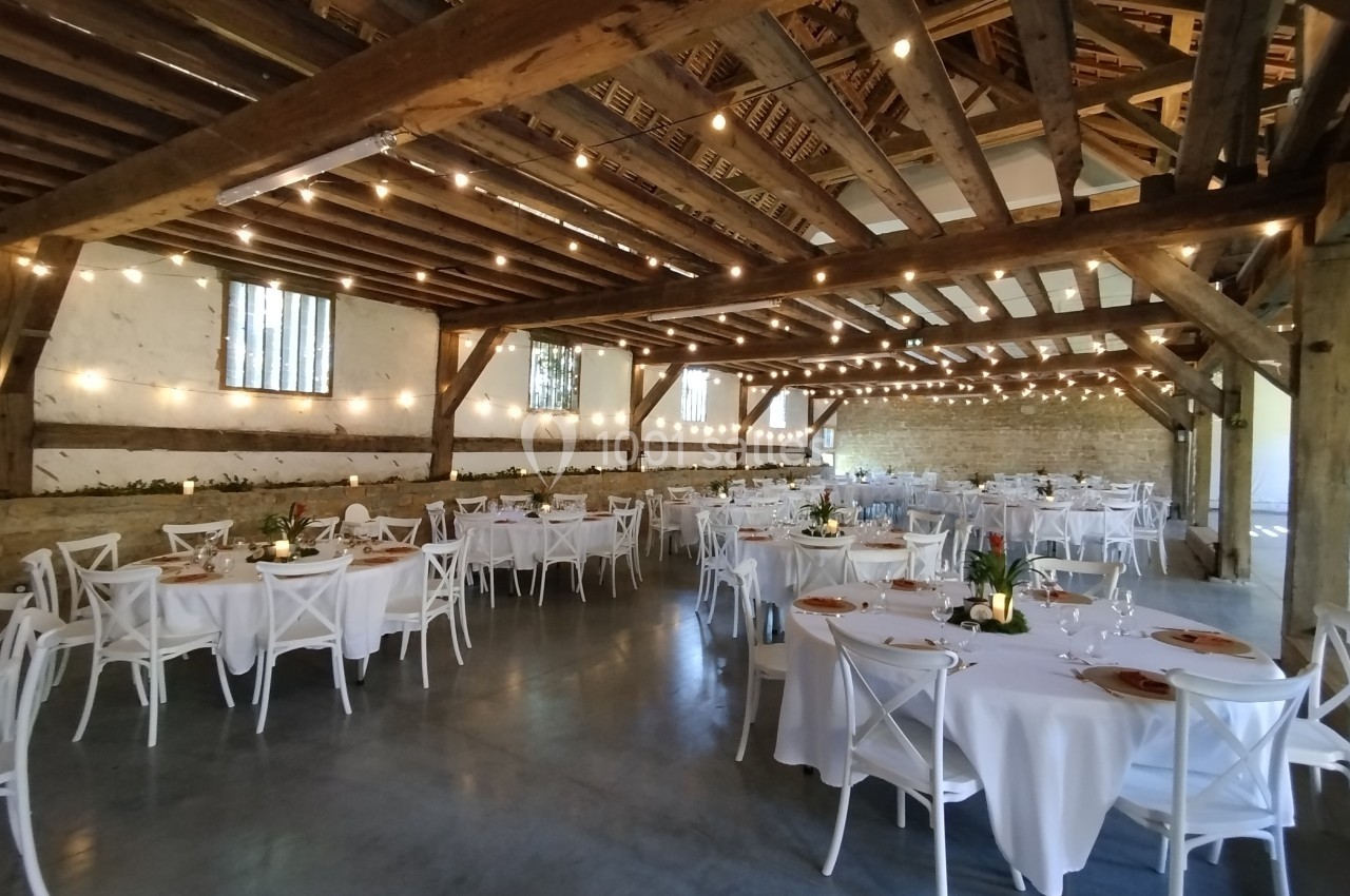 Salle de réception rustique avec tables rondes dressées, chaises blanches et guirlandes lumineuses suspendues.