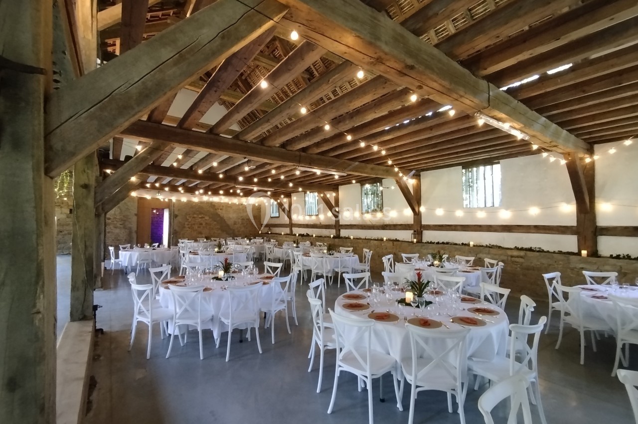 Salle de réception rustique avec poutres apparentes, tables rondes dressées et guirlandes lumineuses suspendues.