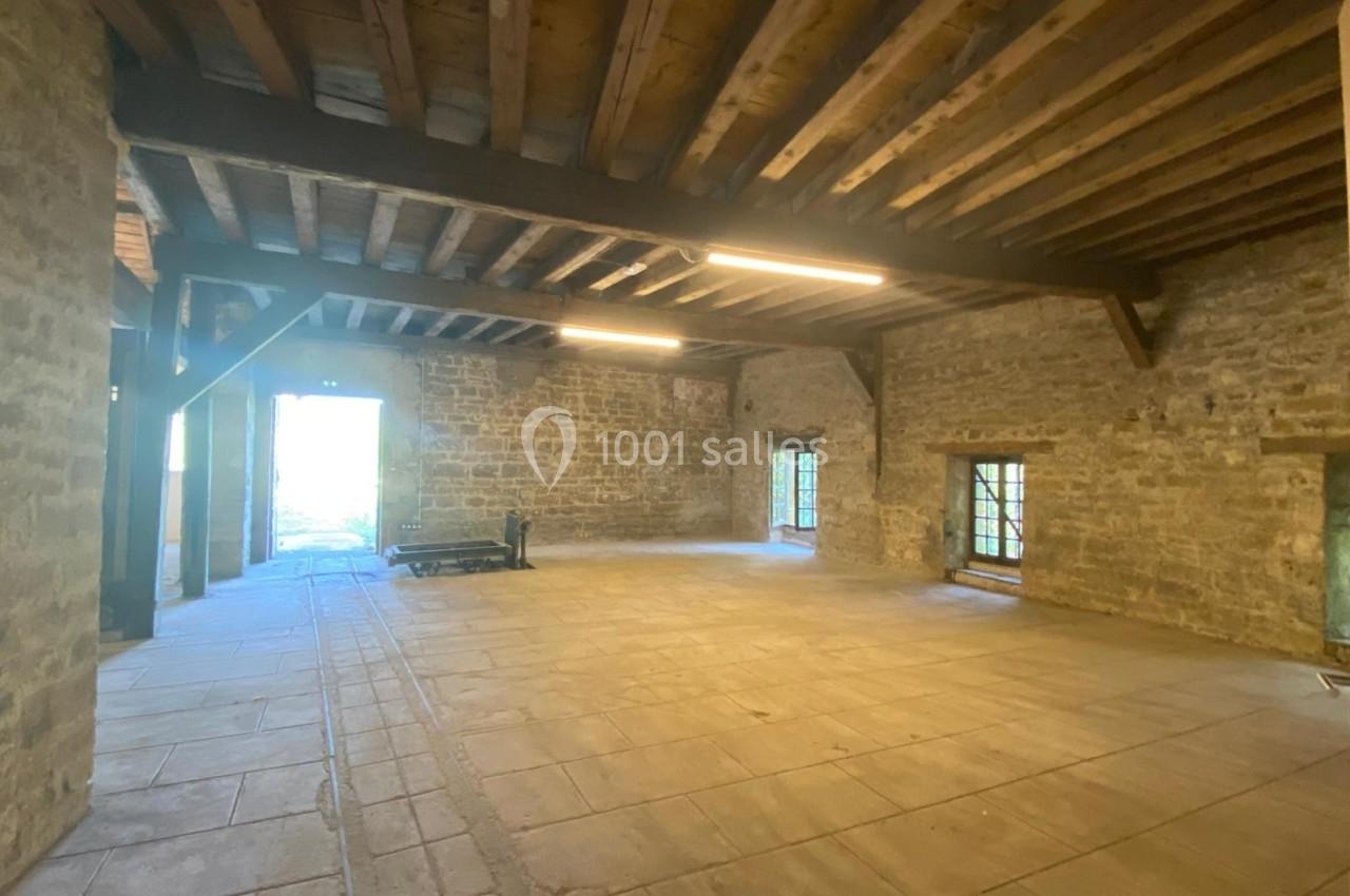 Intérieur d'une grande pièce en pierre avec plafond en poutres apparentes, sol en dalles et lumière naturelle.