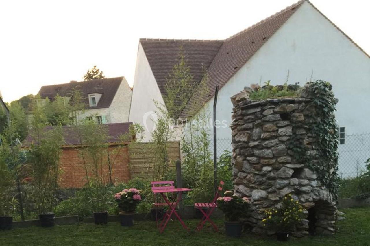 Petit jardin avec une table et des chaises roses, entouré de plantes et d'un puits en pierre devant des maisons.