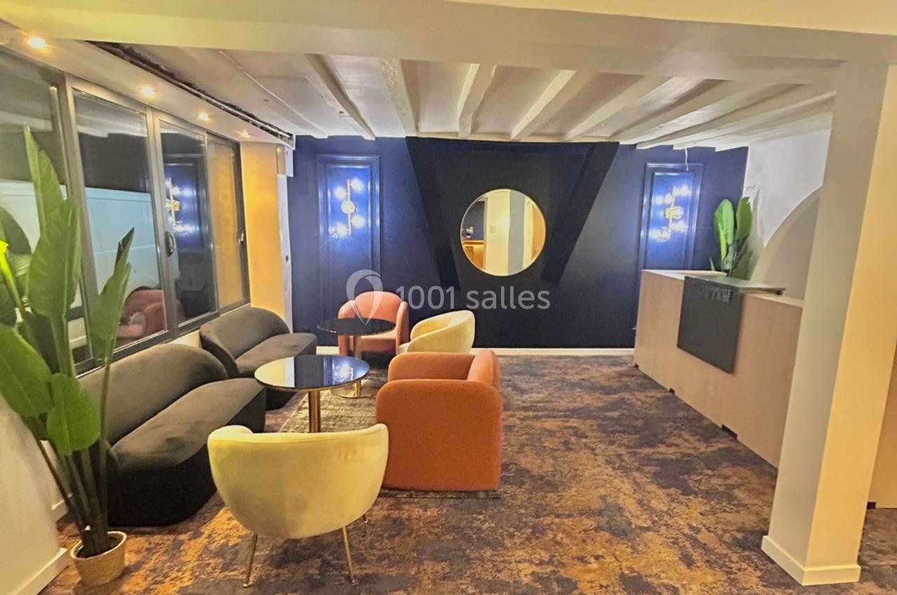 Espace lounge moderne avec fauteuils colorés, table basse, miroir rond et plantes, éclairé par des suspensions.