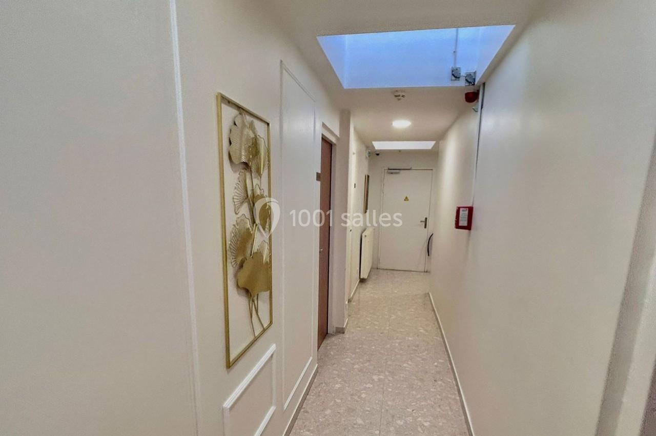 Couloir lumineux avec murs blancs, tableau floral encadré à gauche, porte au fond et éclairage naturel par une lucarne.