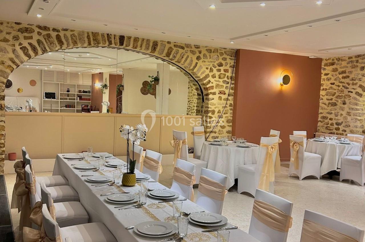 Salle de réception élégante avec tables dressées, nappes blanches, chaises décorées et murs en pierre apparente.
