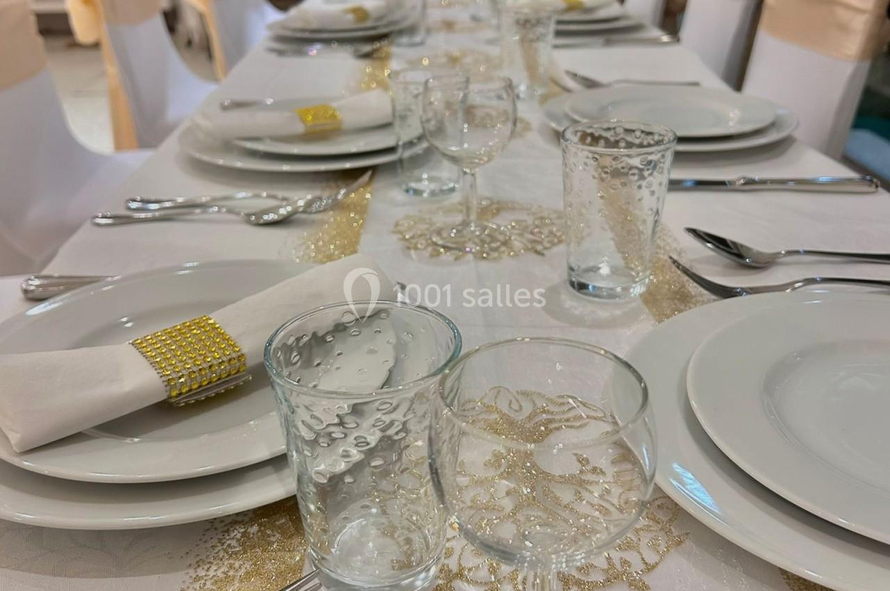 Table dressée avec une nappe blanche et dorée, couverts alignés, assiettes, verres et serviettes décorées.