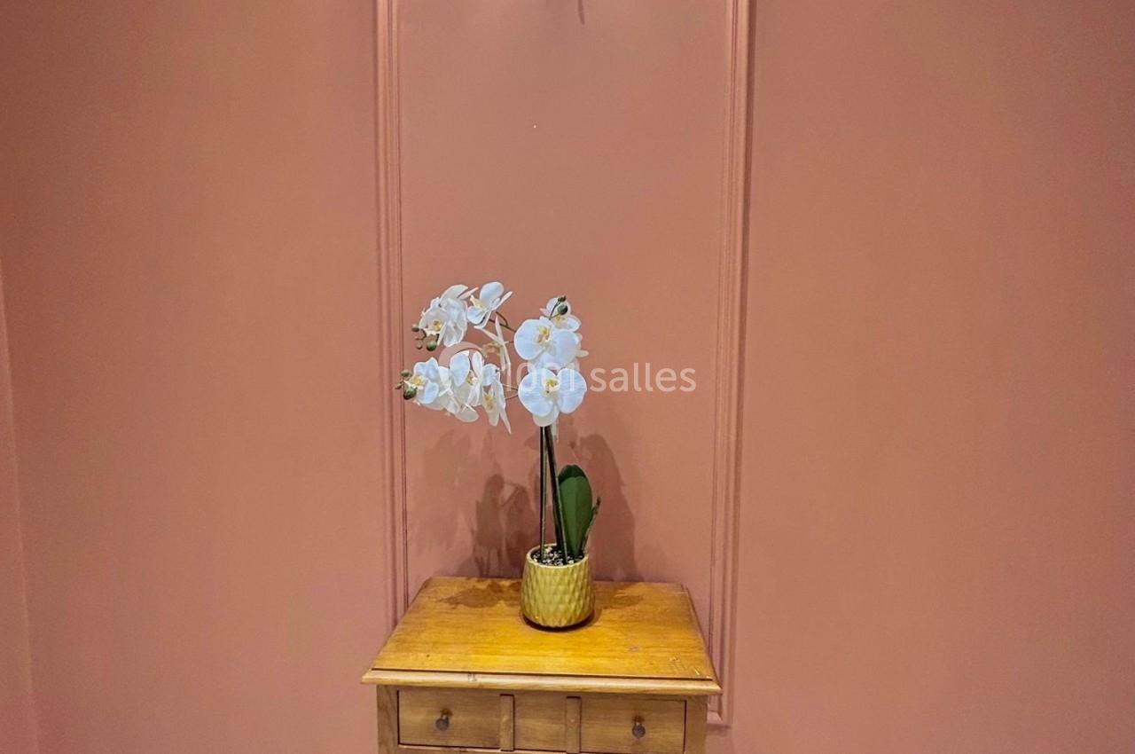 Table en bois avec un vase contenant des orchidées blanches, placée contre un mur rose sous une applique murale allumée.
