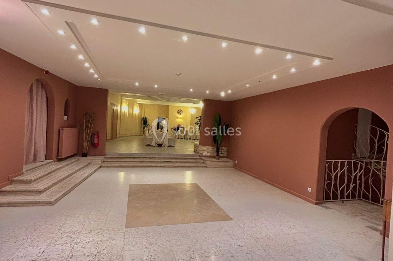 Salle spacieuse avec sol en marbre, murs beige et rouge, éclairage encastré et escalier menant à un espace surélevé.