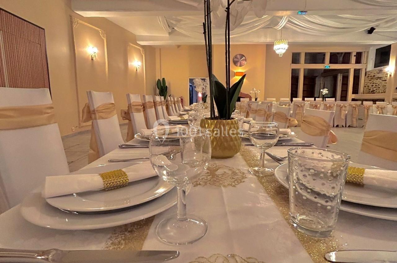 Salle de réception décorée avec des tables dressées, nappes blanches et dorées, et une orchidée en centre de table.