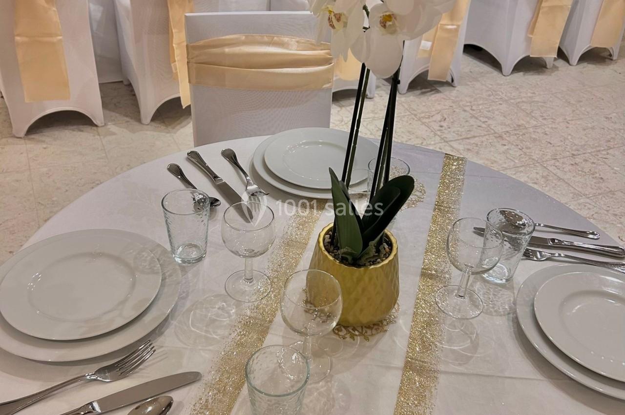 Table dressée avec nappes blanches, orchidée en centre de table, couverts et verres alignés, chaises décorées de nœuds dorés.