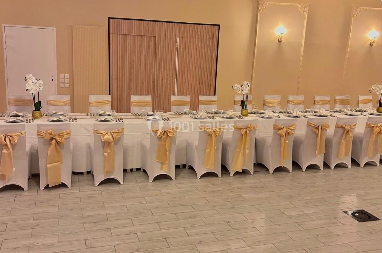 Salle de réception avec une longue table dressée, chaises blanches décorées de nœuds dorés et éclairage chaleureux.