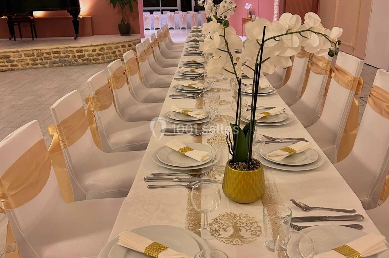 Table élégamment dressée avec nappes blanches, orchidées en centre de table, et chaises décorées de rubans dorés.