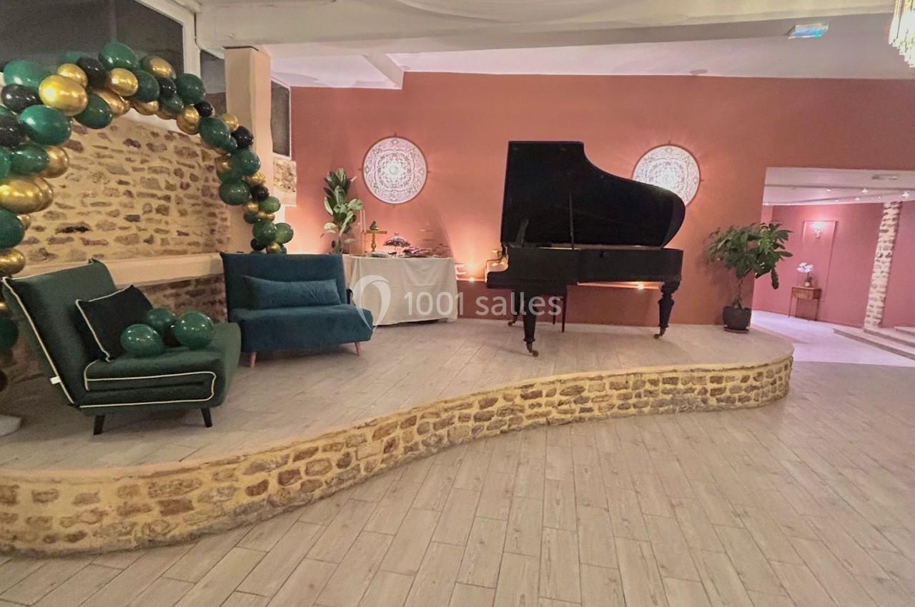 Salle décorée avec un piano à queue noir, un fauteuil bleu, des ballons verts et dorés, et un mur en pierre.
