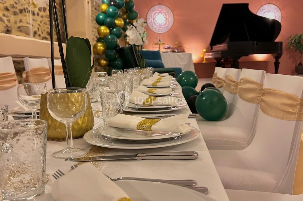 Table élégamment dressée avec vaisselle blanche, décorations dorées et ballons, piano en arrière-plan.