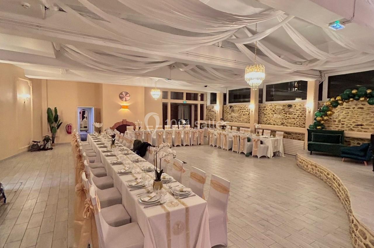 Salle de réception décorée avec des tables alignées, nappes blanches, chaises ornées et éclairage chaleureux.