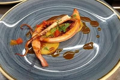 Salle lumineuse avec murs en pierre, mobilier moderne, lampes rouges suspendues et vue sur une entrée vitrée.