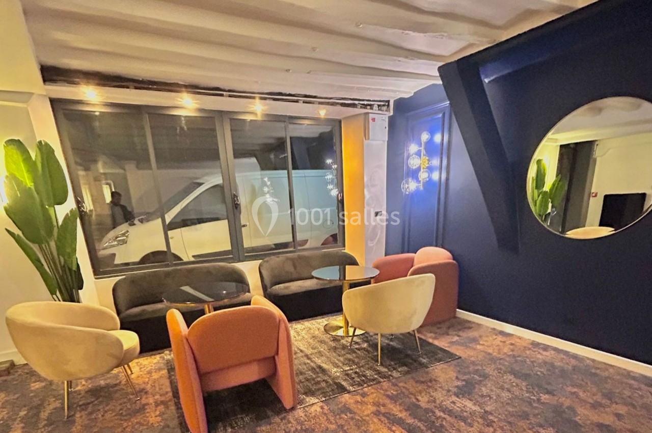 Espace salon avec fauteuils colorés, table basse, miroir rond, mur bleu et grande fenêtre donnant sur l'extérieur.