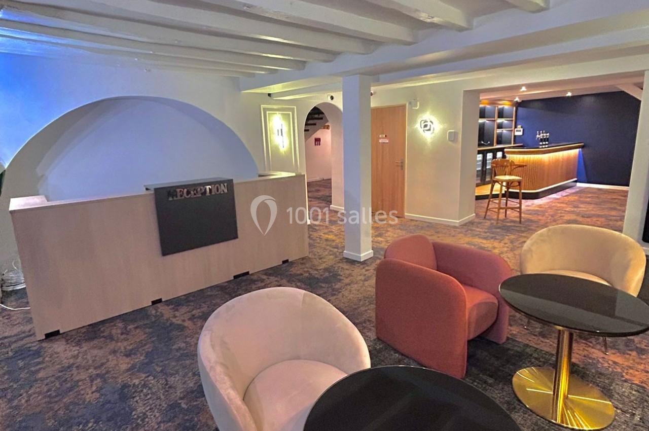 Espace d'accueil avec comptoir de réception, coin salon avec fauteuils et tables, et bar en arrière-plan.