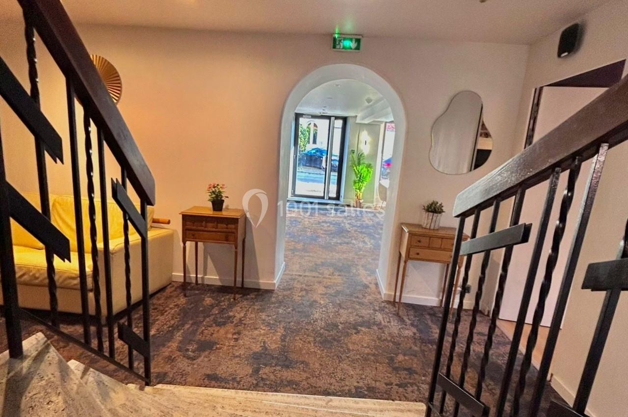 Vue d'un couloir intérieur avec escalier, mobilier en bois, plantes décoratives et porte vitrée donnant sur l'extérieur.