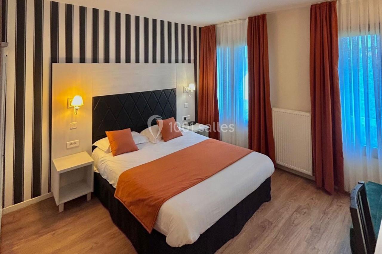 Chambre d'hôtel avec lit double, tête de lit rembourrée, murs à rayures, rideaux orange et éclairage doux.