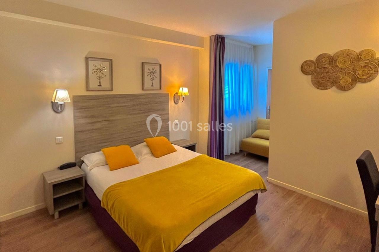 Chambre lumineuse avec lit double, couvre-lit jaune, canapé en arrière-plan et décoration murale.