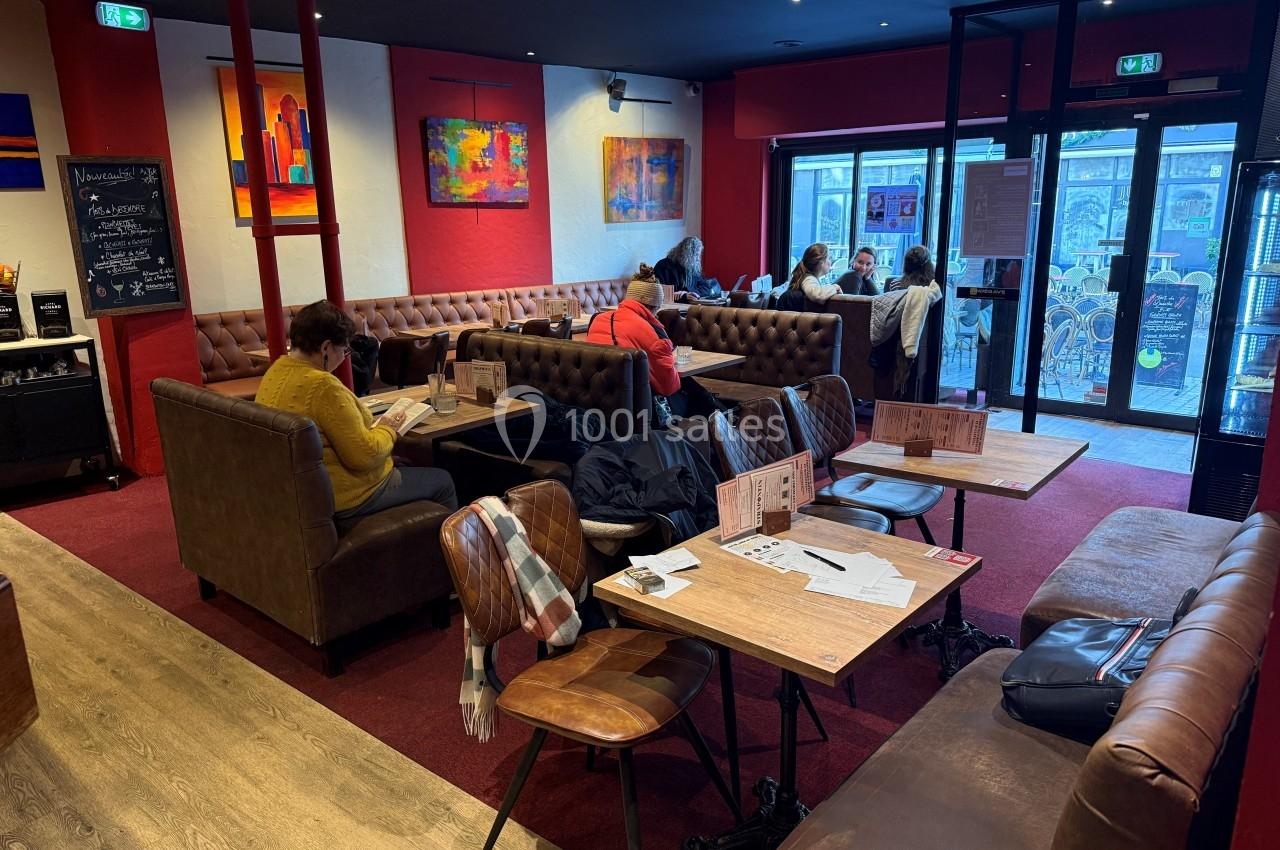 Intérieur d'un café avec des tables en bois, des banquettes et des clients assis, certains lisant ou discutant.